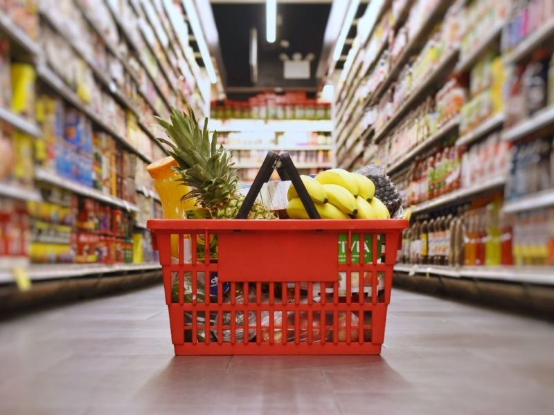 Red shopping basket filled with fresh fruits and vegetables in grocery store aisle.