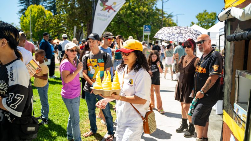 A crowd of people at the Bizerkeley Food Fest with a woman wearing all white waking through the crowd holding Dole Whips as festival goers expolore