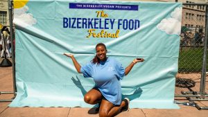 Erika Hazel stands in front of The Bizerkeley Food Festival sign, an aqua blue with orange and navy writing. Erika Hazel kneels down, wearing a light blue festival shirt with her arms in the air.
