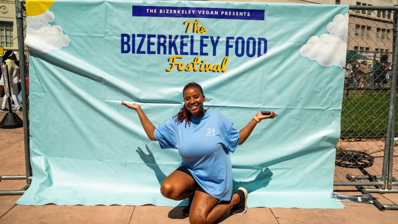 Erika Hazel stands in front of The Bizerkeley Food Festival sign, an aqua blue with orange and navy writing. Erika Hazel kneels down, wearing a light blue festival shirt with her arms in the air.