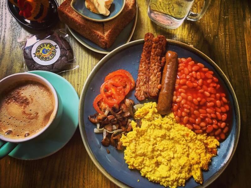 A plate with vibrant yellow tofu scramble, orange baked beans. brown sausage and tempeh and grilled tomatoes and mushrooms sits on a table next to a mug of coffee in London