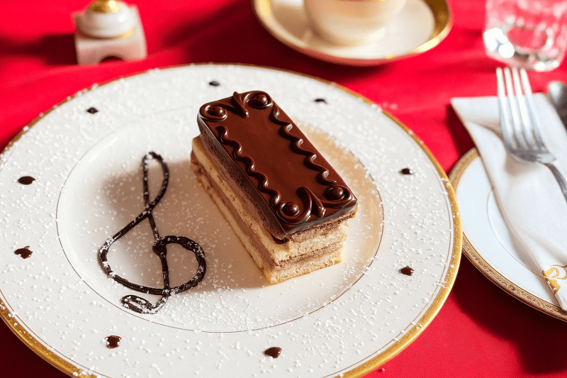 A rectangular slice of vegan opera cake topped with glossy chocolate ganache and intricate decorative swirls, served on an elegant white plate with gold trim. The plate is dusted with powdered sugar and features a chocolate treble clef design beside the dessert.